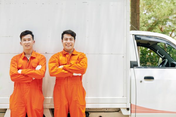 Positive confident Asian movers in orange uniform standing in front of van with arms crossed and smiling at camera
